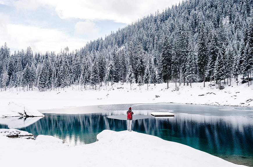 Hiker with Red Backpack Standing by Frozen Alpine Lake in Winter