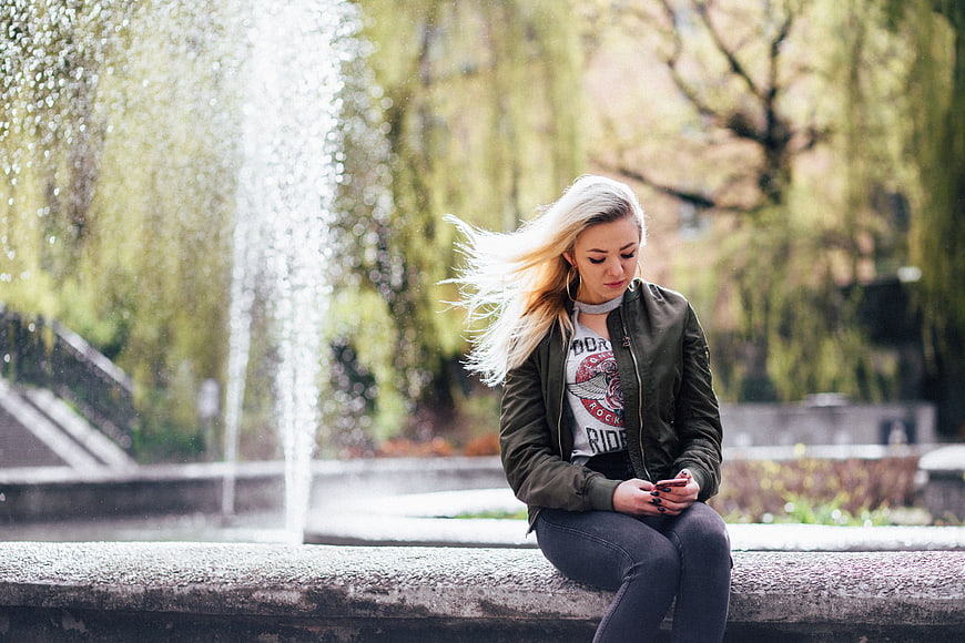 Young Blonde Woman Sitting by Fountain Using Smartphone