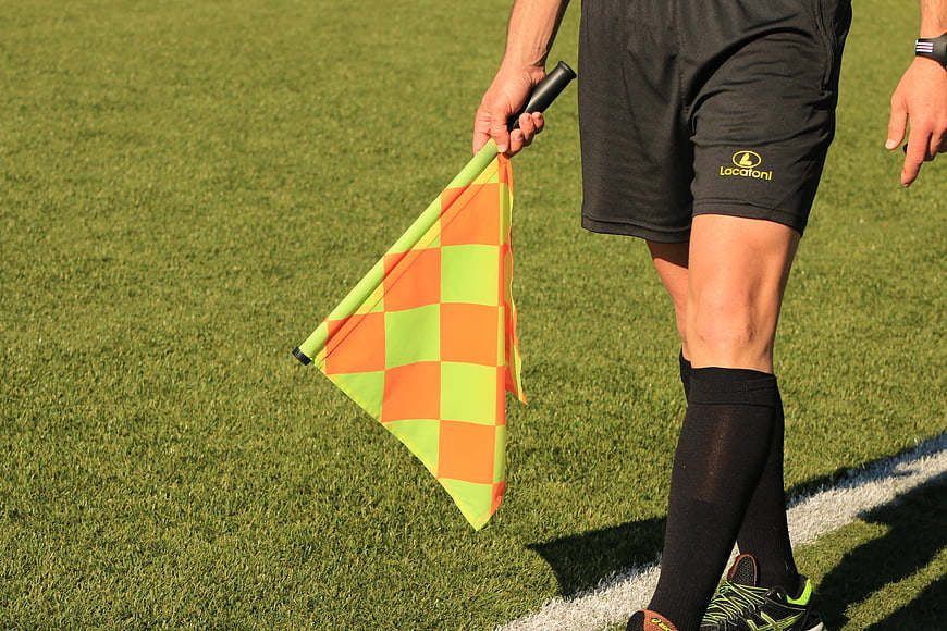 Soccer Referee Holding Orange And Green Checkered Flag On Grass Field