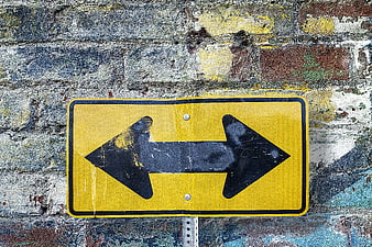 Yellow Two-Way Arrow Sign on Weathered Brick Wall
