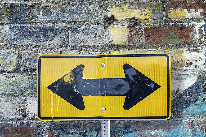 Yellow Two-Way Arrow Sign on Weathered Brick Wall