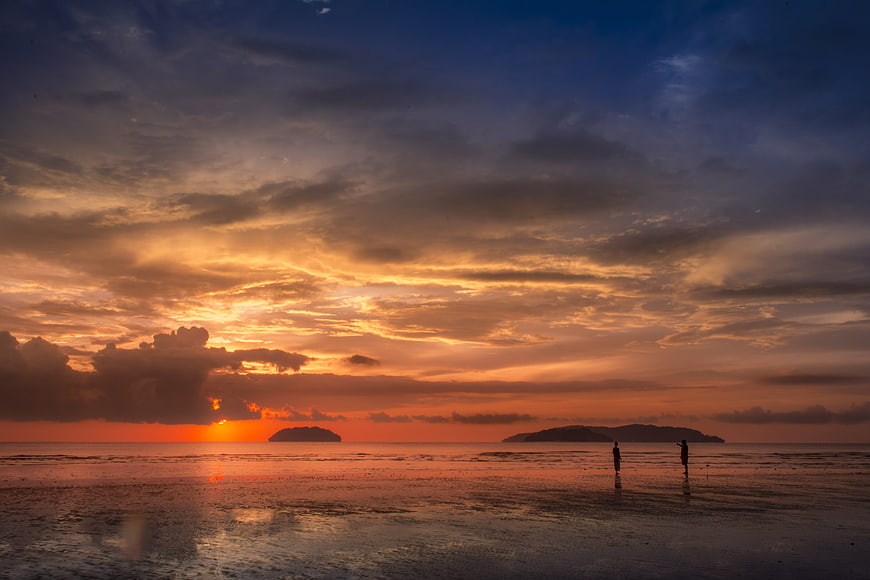 Dramatic Sunset Over Beach With Silhouetted Figures And Distant Islands