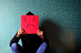 Person holding red sad face paper mask against blue wall