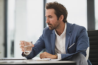 Thoughtful businessman in blue blazer holding water glass at office desk