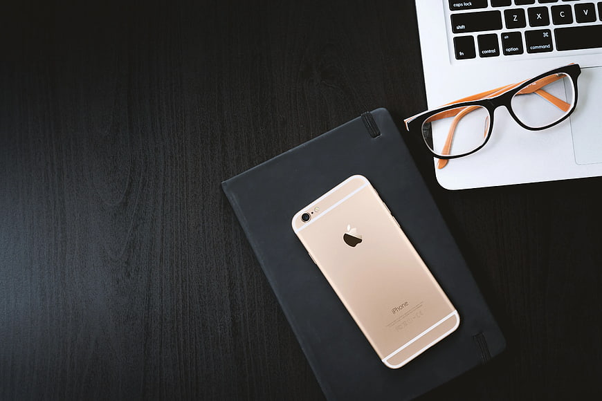 Flat lay of gold iPhone, black notebook, and laptop with glasses