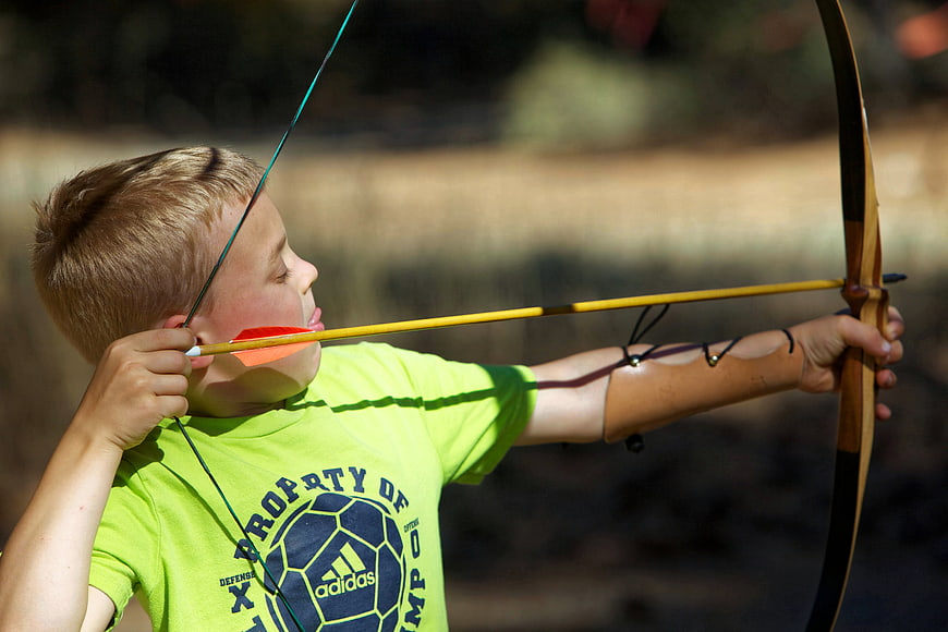 Young boy aiming a traditional wooden bow and arrow outdoors