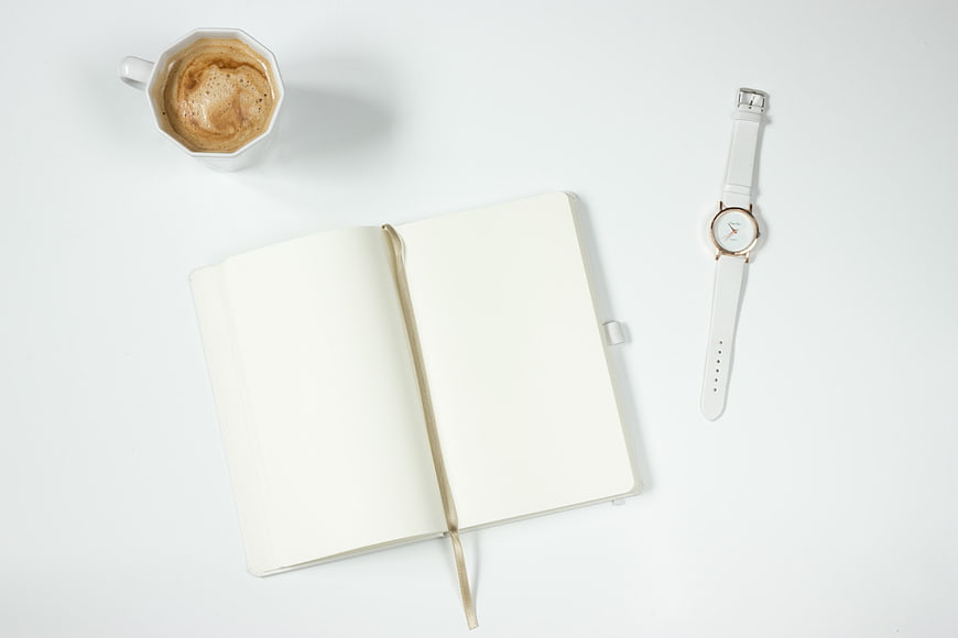 White notebook, coffee cup, and wristwatch on a clean white desk surface