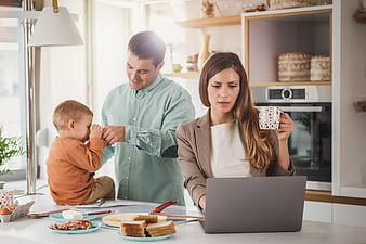 Busy mother working on laptop while father plays with toddler at breakfast