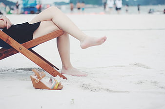 Woman relaxing barefoot on a wooden beach chair with sandals on the sand