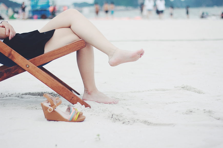Woman relaxing barefoot on a wooden beach chair with sandals on the sand