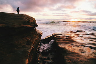 Lone Figure Standing on Rocky Cliff Overlooking Ocean Sunset