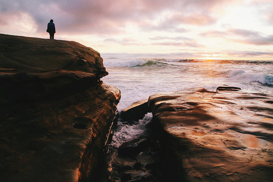 Lone Figure Standing on Rocky Cliff Overlooking Ocean Sunset