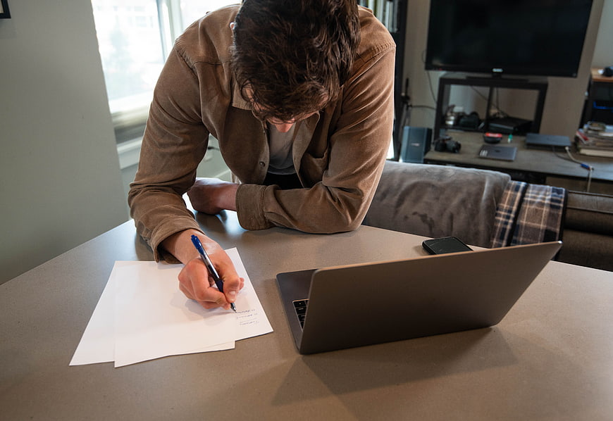 Man writing on paper at desk with laptop