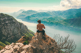 Shirtless Man Sitting on Cliff Edge Overlooking Mountain Lake