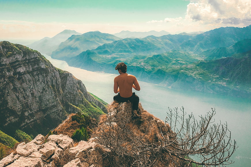 Shirtless Man Sitting on Cliff Edge Overlooking Mountain Lake