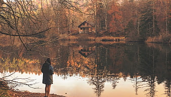 Woman standing by lake with cabin reflection in autumn forest