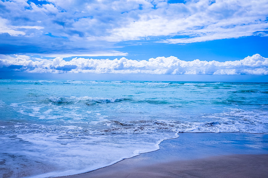 Vibrant Blue Ocean Waves Crashing on Sandy Beach Under Cloudy Sky