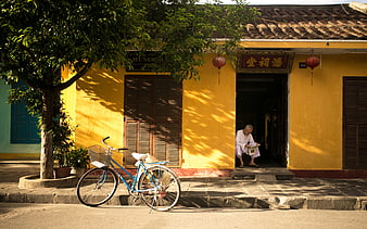 Old Man Reading Newspaper in Front of Yellow House with Bicycle