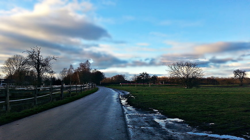 Wet country road winding through green fields under cloudy sky