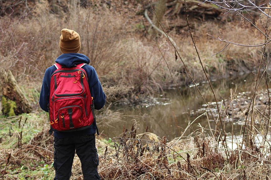 Hiker with Red Backpack Standing by Stream in Woods