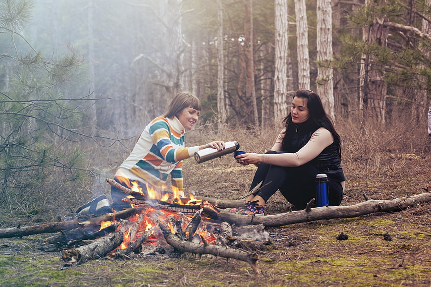 Two women camping and drinking by a bonfire in the forest