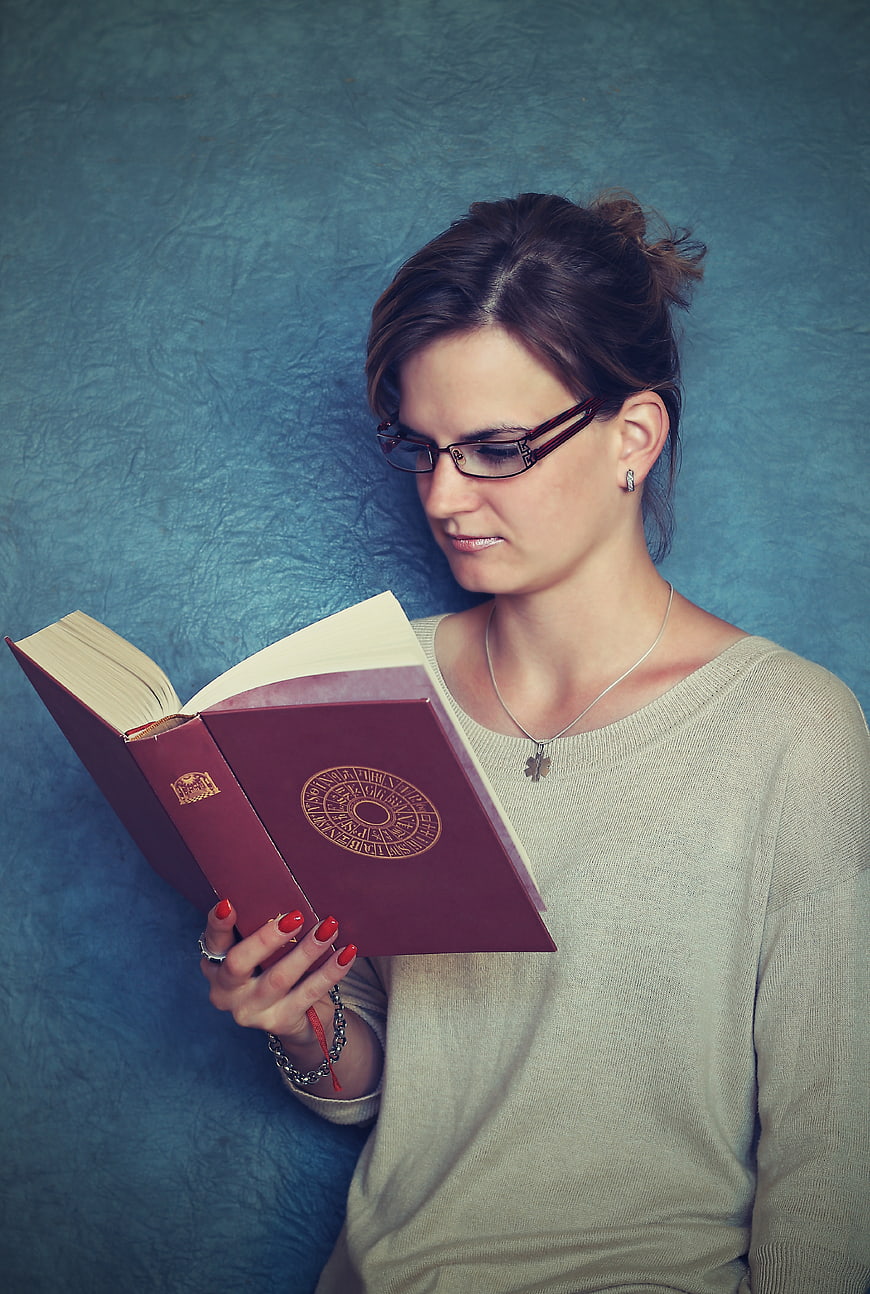 Young woman with glasses reading a red hardcover book against a blue background
