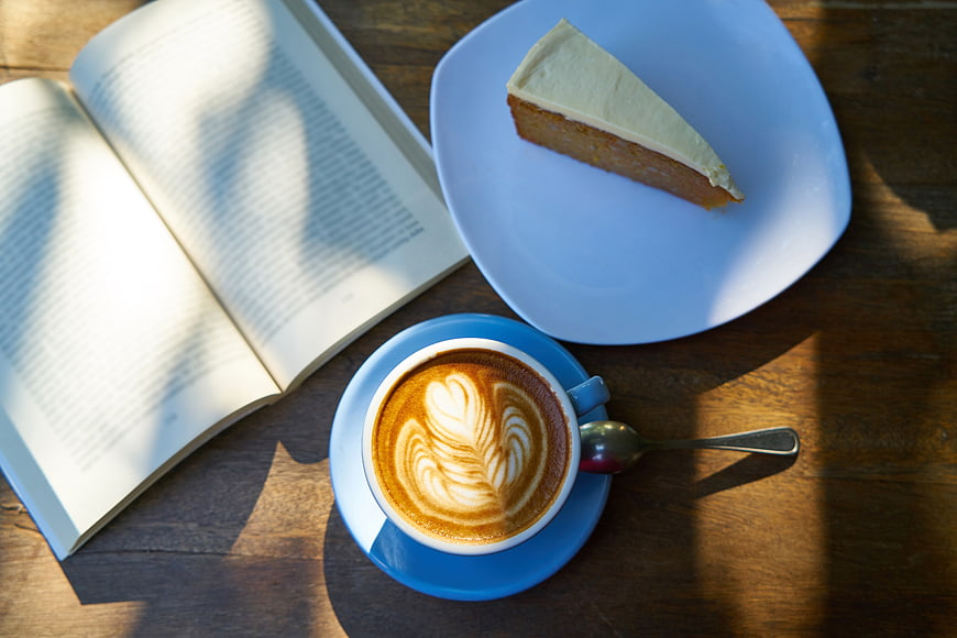 Flat lay of coffee latte art, cake slice, and open book on wooden table