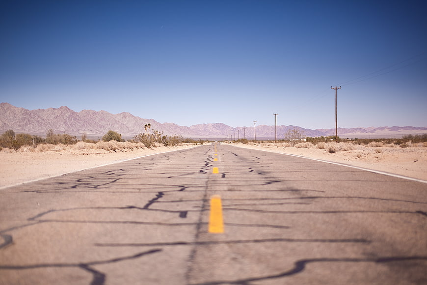 Cracked Desert Highway Leading To Distant Mountains Under Blue Sky