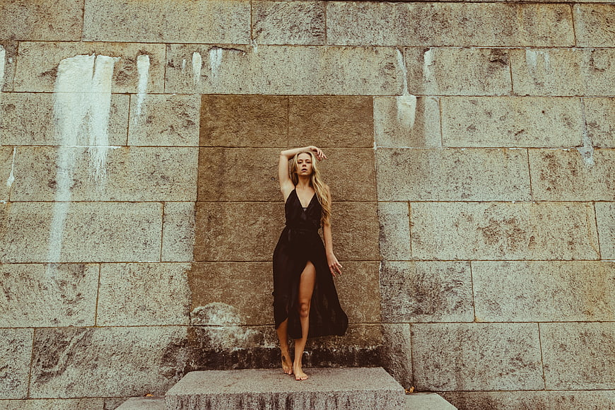 Blonde woman in black dress posing against textured stone wall