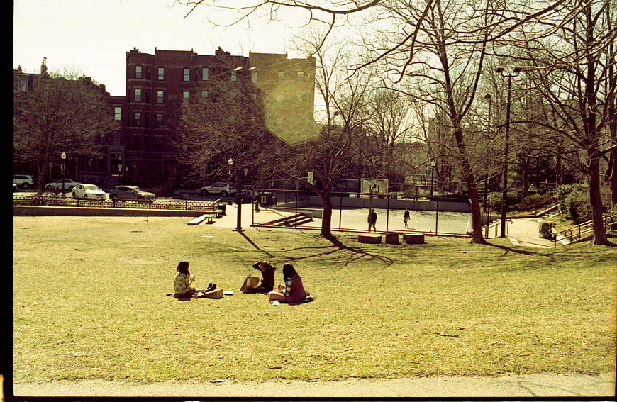 People Relaxing On Grass In Urban City Park With Apartment Buildings