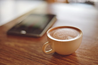 Close-up of frothy latte on wooden table with blurred smartphone