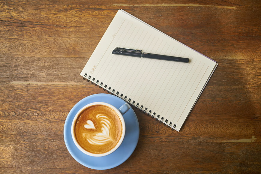 Flat lay of coffee with latte art, notebook, and pen on wooden table