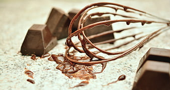 Melted chocolate on a whisk with dark chocolate blocks on a textured surface