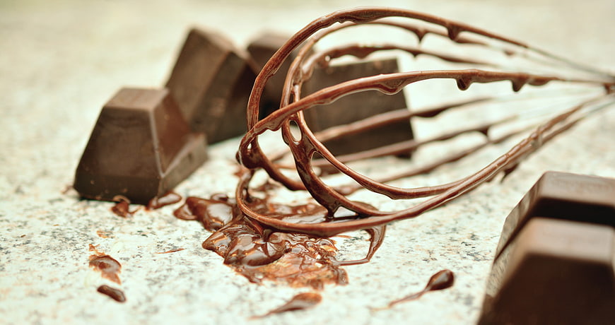 Melted chocolate on a whisk with dark chocolate blocks on a textured surface
