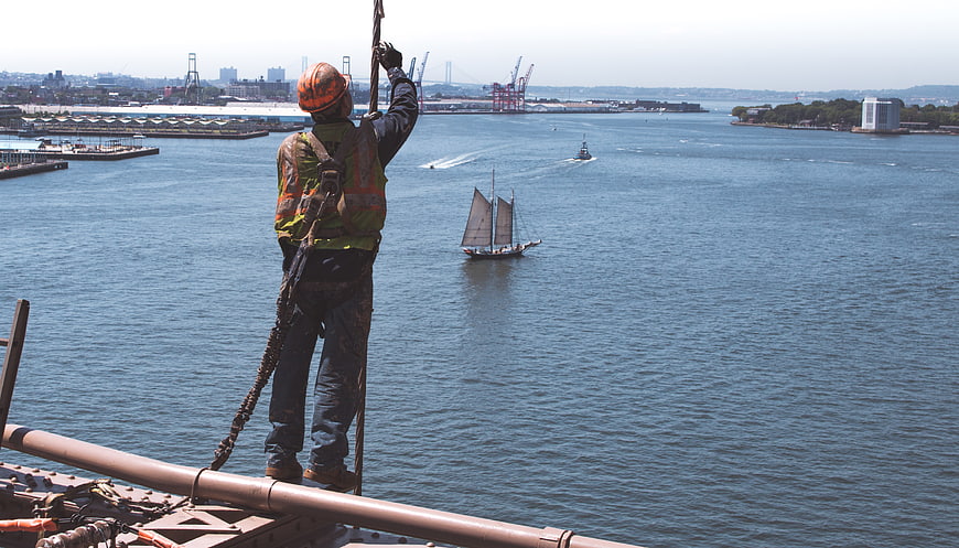 Construction worker on bridge overlooking harbor with sailboat