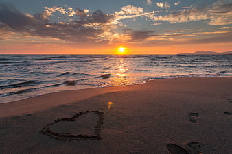 Heart drawn in sand on beach at sunset with footprints