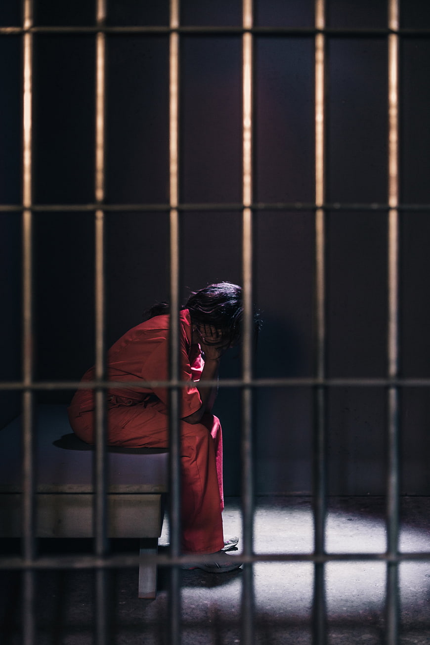 Sad woman in orange jumpsuit sitting in prison cell