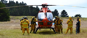 Firefighters in yellow gear surrounding red rescue helicopter in grassy field