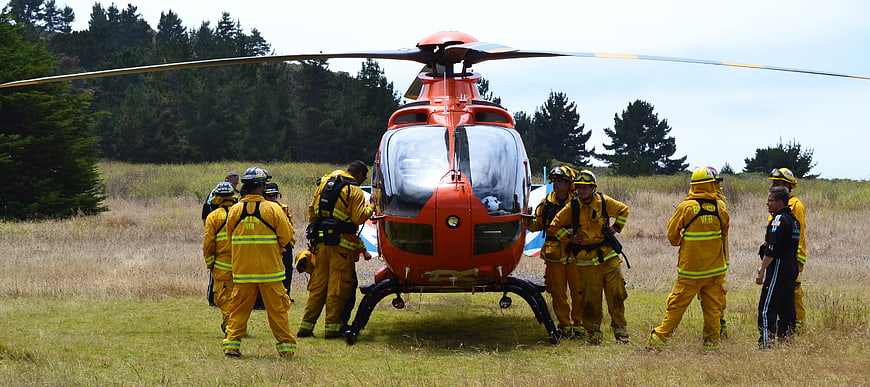 Firefighters in yellow gear surrounding red rescue helicopter in grassy field