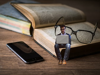 Miniature businessman sitting on book with laptop and glasses