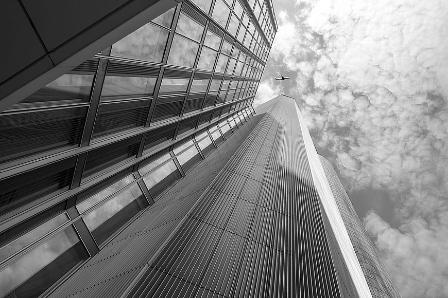 Black and white low-angle view of modern skyscrapers with airplane in cloudy sky