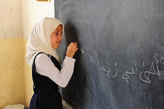 Young girl in hijab writing Arabic script on a classroom blackboard