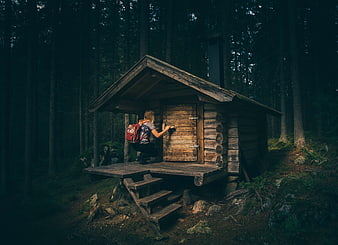 Woman with backpack opening door of rustic log cabin in dark forest
