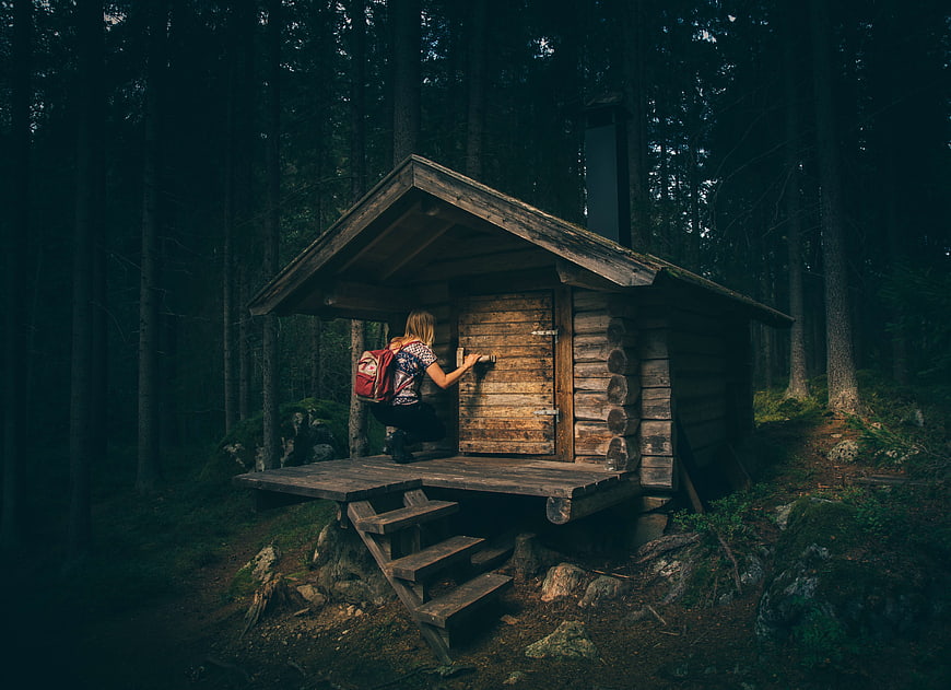Woman with backpack opening door of rustic log cabin in dark forest