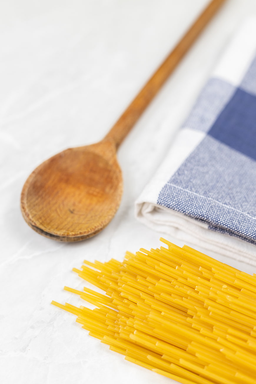 Uncooked spaghetti, wooden spoon, and blue checkered napkin on white marble