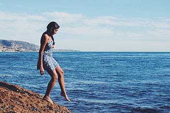 Woman in Blue Dress Walking on Coastal Rocks by Ocean