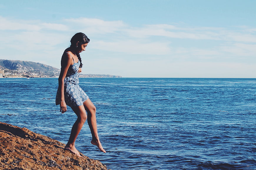 Woman in Blue Dress Walking on Coastal Rocks by Ocean