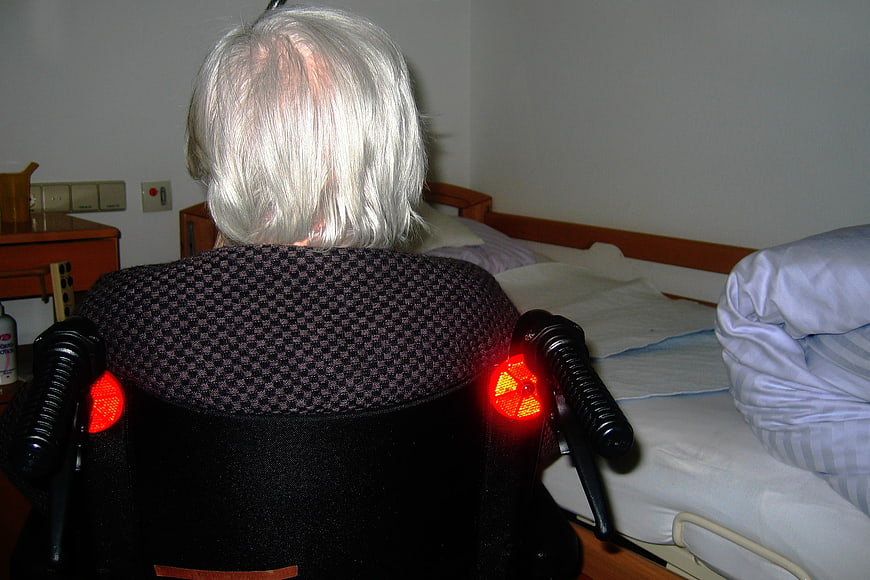 Senior woman with grey hair sitting in wheelchair facing bed