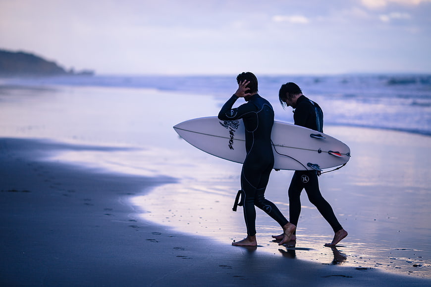 Two male surfers in wetsuits walking on a blue beach at dusk