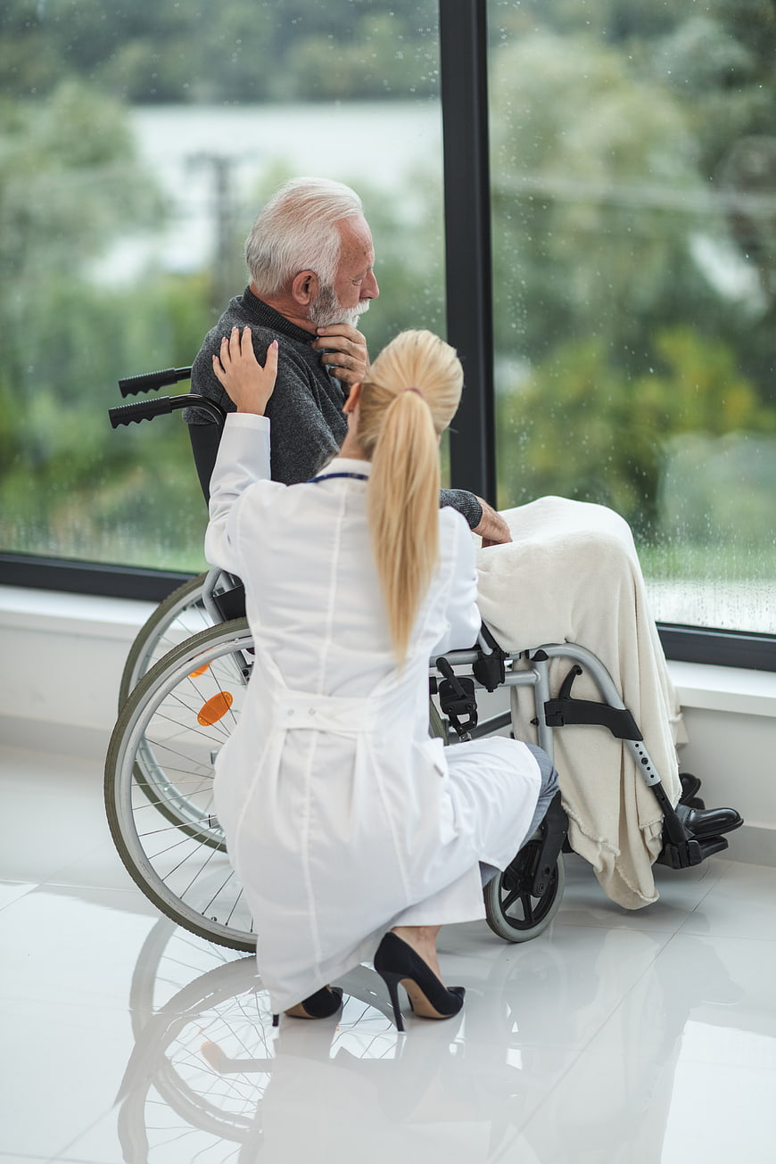 Female nurse comforting elderly man in wheelchair by window
